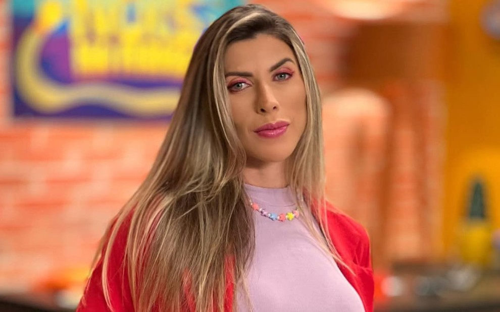 Ana Paula Minerato usa uma blusa lilás e um casaco laranja; ela encara a câmera, séria