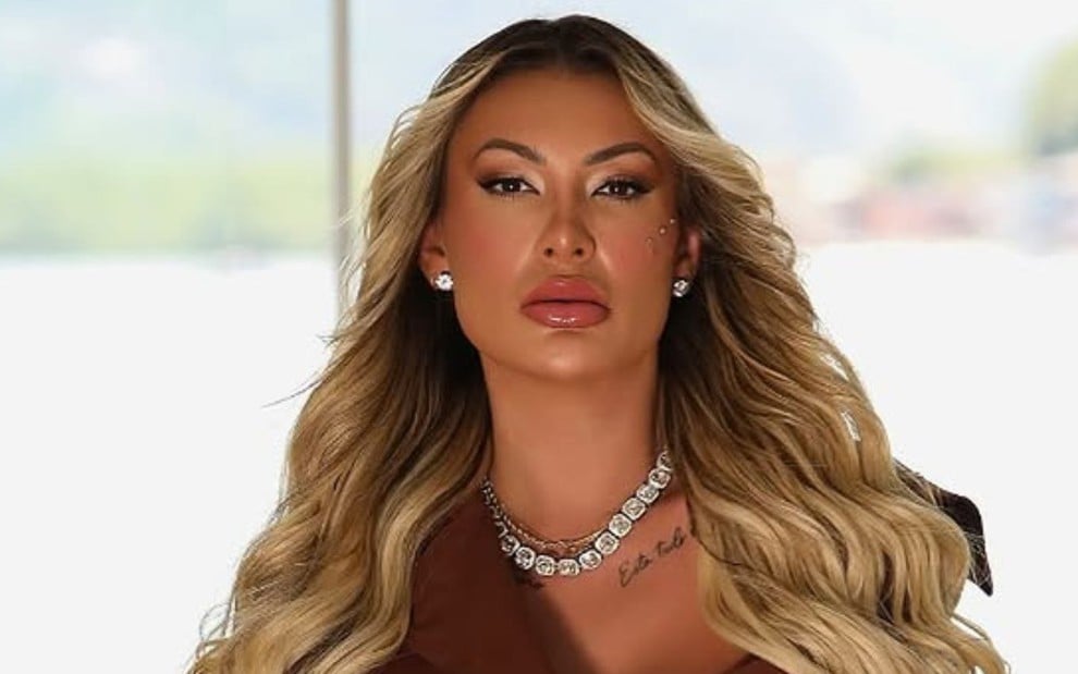Andressa Urach sentada em frente a uma janela, com feição séria, usando joias e cabelo loiro
