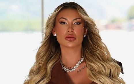 Andressa Urach sentada em frente a uma janela, com feição séria, usando joias e cabelo loiro