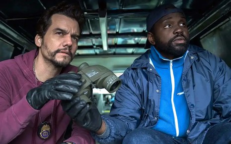 Wagner Moura usa um moletom roxo e segura binóculos em cena de Ladrões de Drogas; ao lado dele está o ator Brian Tyree Henry