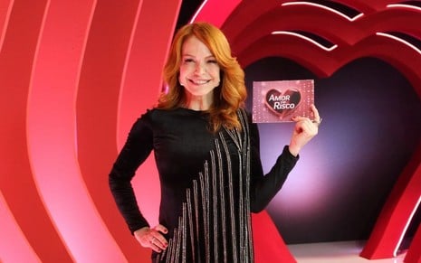 mulher ruiva posa para foto em estúdio de TV; ela usa um vestido preto com detalhes brilhantes