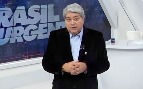 Homem apresentando programa de TV em estúdio, com letreiro "Brasil Urgente" ao fundo. Ele veste paletó preto e camisa azul.