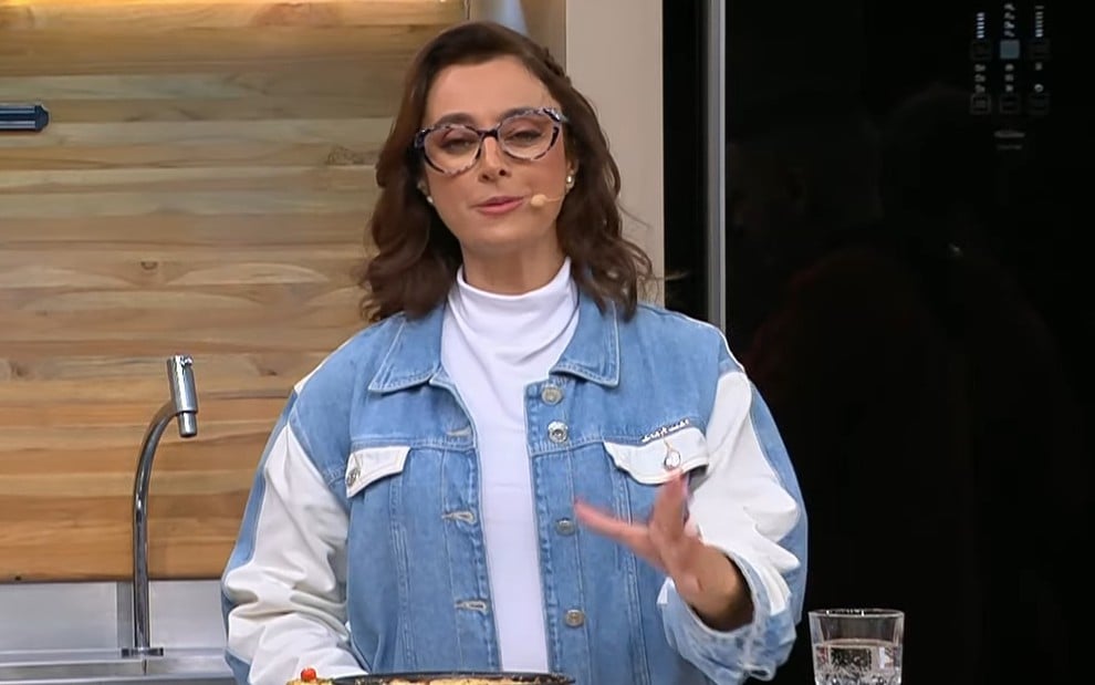 Mulher de cabelos castanhos ondulados usa óculos de aro grosso, blusa branca de gola alta e jaqueta jeans bicolor.