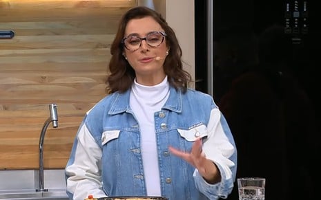 Mulher de cabelos castanhos ondulados usa óculos de aro grosso, blusa branca de gola alta e jaqueta jeans bicolor.