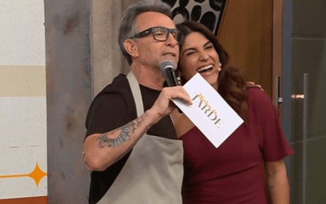 Homem de cabelo grisalho, óculos escuros e avental claro abraça mulher de vestido vinho; ele segura microfone e cartão com o texto “Melhor da Tarde”.