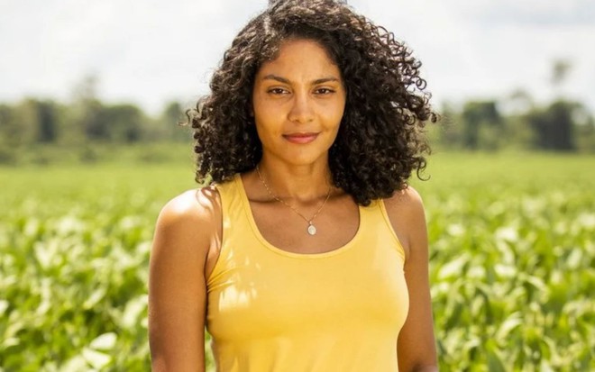 Bárbara Reis caracterizada como Aline em Terra e Paixão (2023), usando regata amarela em um fundo com bastante vegetação