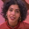 Mulher de cabelo curto e cacheado, veste blusa de tom terroso com gola fechada e usa brincos dourados grandes em formato de gota.