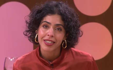 Mulher de cabelo curto e cacheado, veste blusa de tom terroso com gola fechada e usa brincos dourados grandes em formato de gota.