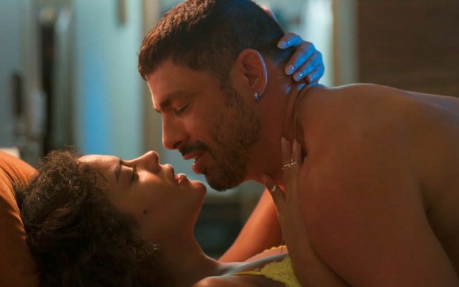 Bella Campos e Cauã Reymond deitados em cama em cena no primeiro capítulo do remake de Vale Tudo