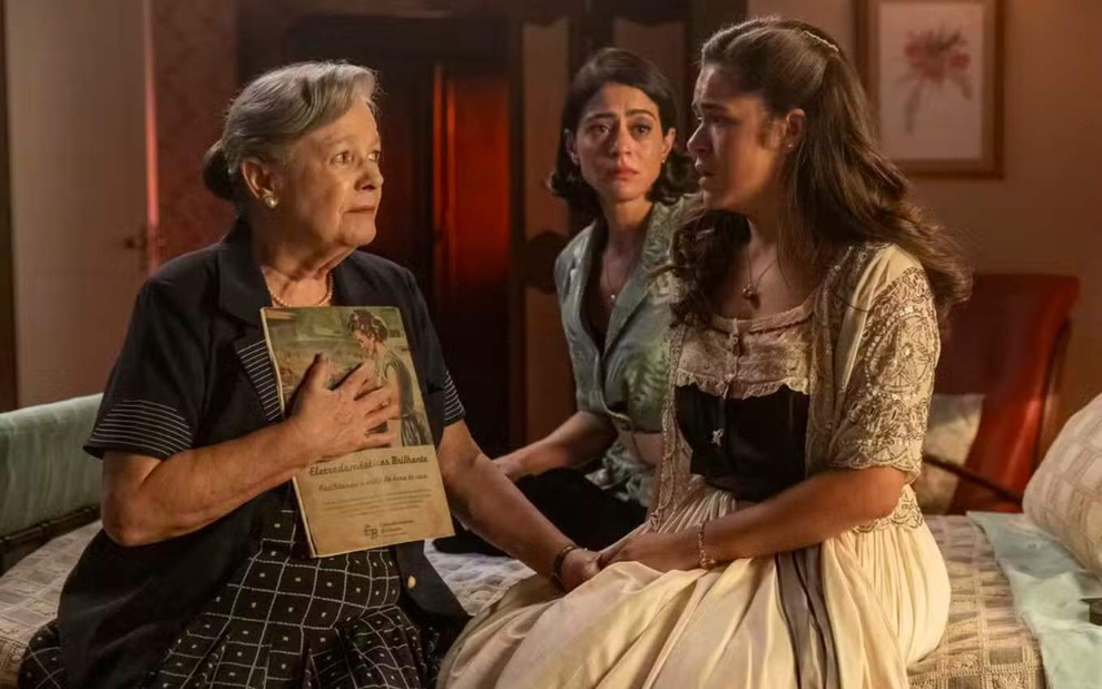 As atrizes Bete Mendes e Carol Castro olhando para a atriz Maisa, as três sérias sentadas numa cama, em cena de Garota do Momento