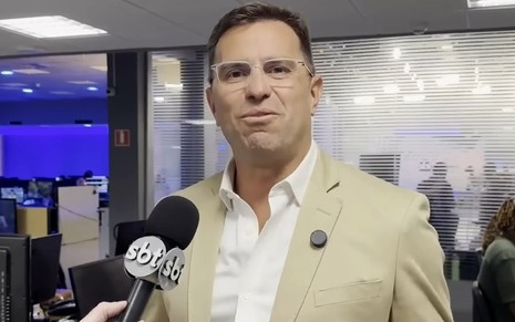 O jornalista Rodrigo Bocardi, de blazer bege, camisa branca e óculos de armação transparente, concede entrevista em uma redação diante de um microfone do SBT.