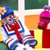 Imagem colorida mostra os palhaços Patati e Patatá sentados sorrindo em um sofá, com um fantoche rosa ao fundo em cenário infantil alegre