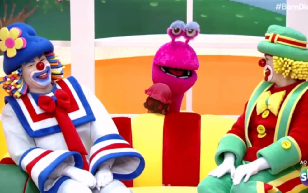 Imagem colorida mostra os palhaços Patati e Patatá sentados sorrindo em um sofá, com um fantoche rosa ao fundo em cenário infantil alegre
