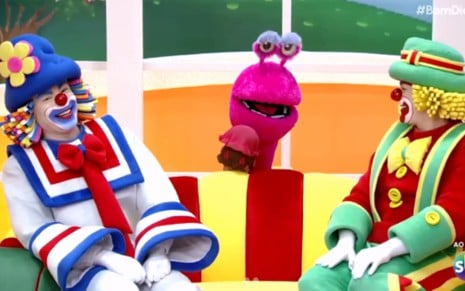 Imagem colorida mostra os palhaços Patati e Patatá sentados sorrindo em um sofá, com um fantoche rosa ao fundo em cenário infantil alegre