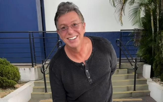 Boninho veste uma blusa de manga longa preta, está sorrindo e usa óculos de grau