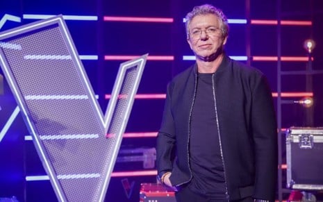 homem grisalho posa para foto em estúdio de reality show; ele veste roupas pretas