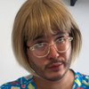Montagem lado a lado mostra um homem com barba rala e óculos; à direita, ele aparece com peruca loira e jaleco estampado.