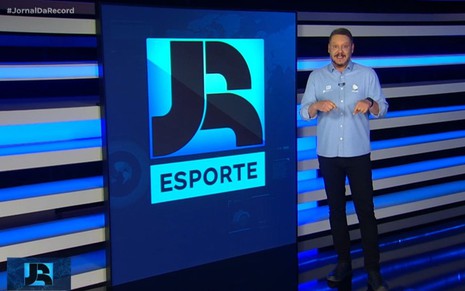 Bruno Laurence está em pé em frente a telão com o logo do JR Esporte