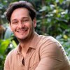 Bruno Luperi usa uma camisa marrom e sorri; ao fundo há várias plantas