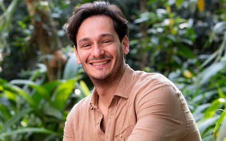 Bruno Luperi usa uma camisa marrom e sorri; ao fundo há várias plantas