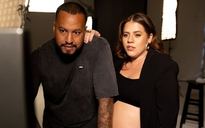Bruno Monteiro e Tati Machado lado a lado em um estúdio fotográfico, os dois usam roupas pretas e olham para uma tela; ela segura no ombro dele