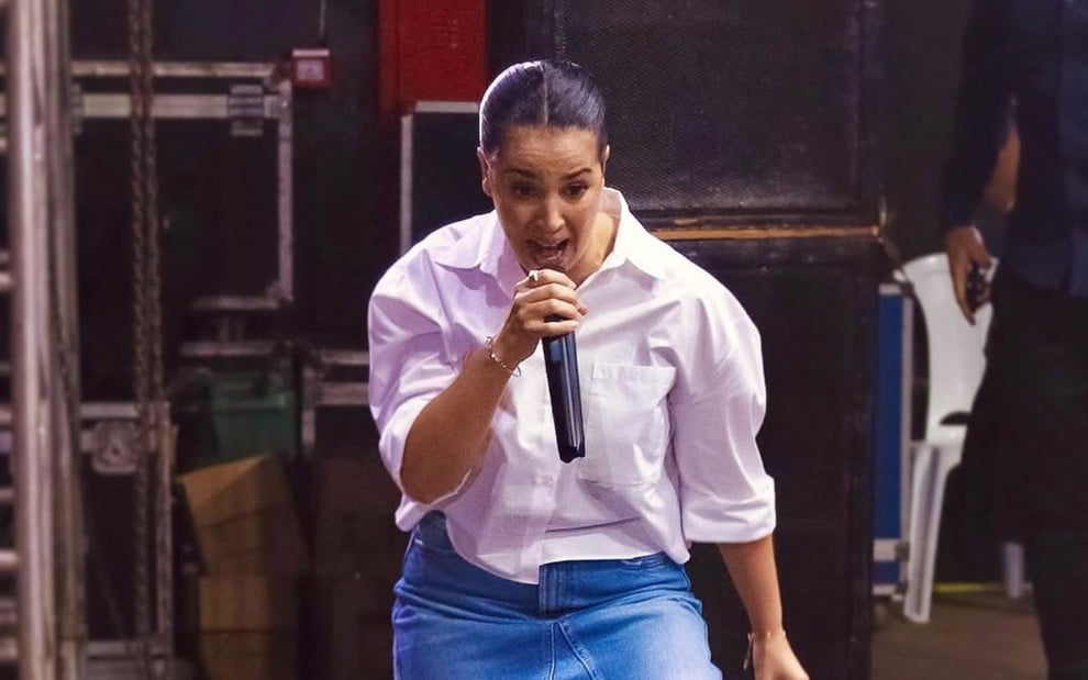 Uma mulher cantando ao microfone em um palco, vestindo camisa branca e saia jeans, com expressão intensa e cabelos presos.