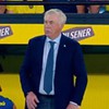 Técnico de terno azul e gravata azul claro em frente a placa amarela da Pilsener durante partida de futebol.