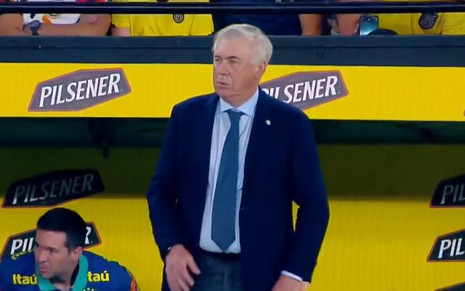 Técnico de terno azul e gravata azul claro em frente a placa amarela da Pilsener durante partida de futebol.