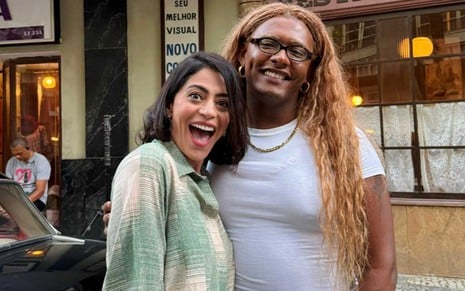 A atriz Carol Castro com expressão muito feliz ao lado da cantora Liniker na cidade cenográfica de Garota do Momento