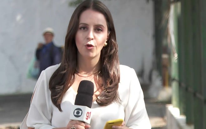 Carol Ianelli no Bom Dia Brasil, de camiseta branca e segurando o microfone da Globo, enquanto homem a encara à distância