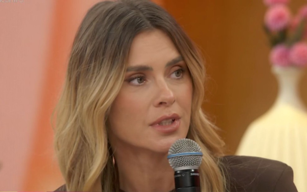 Carolina Dieckmann no sofá do Encontro, com o microfone na mão, usando blazer marrom