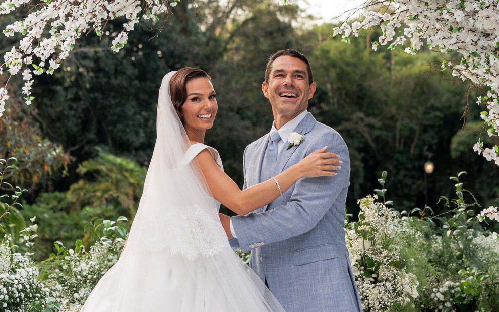 Vestida de noiva, Isis Valverde posa sorridente enquanto segura o braço de Marcus Buaiz, que veste um terno azul claro