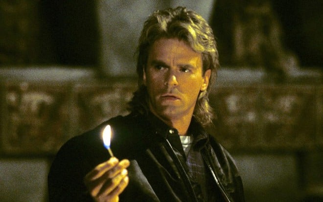 Richard Dean Anderson segura um fósforo aceso em cena da série MacGyver - Profissão: Perigo; ele tem mullet no cabelo, com visual típico dos anos 1980