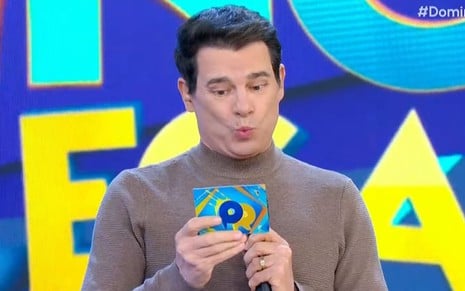 Celso Portiolli faz biquinho enquanto segura microfone e uma ficha no Passa ou Repassa
