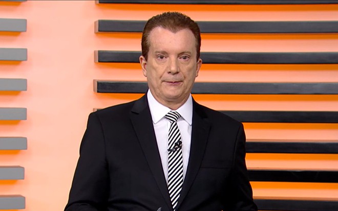 Homem de terno preto e gravata listrada apresenta programa diante de parede com listras horizontais pretas e laranja.