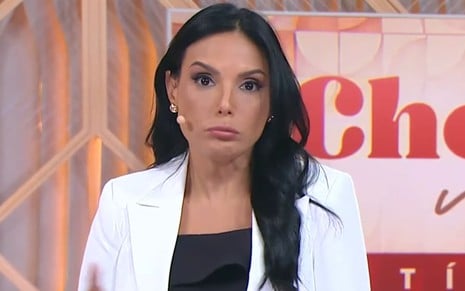 Mulher de cabelos escuros, usando blazer branco e microfone auricular, faz expressão de surpresa em um cenário com fundo colorido