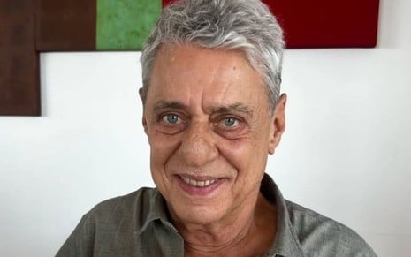 Chico Buarque sorridente, em fundo branco, com um quadro verde e vermelho atrás; ele usa camisa cinza