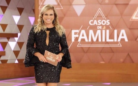 Christina Rocha posando em frente a um telão com o logo do Casos de Família; ela aparece sorridente, com vestido preto de lantejoulas, e segura ficha do programa nas mãos