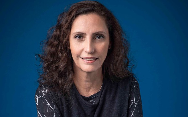 Claudia Souto, a autora de Volta por Cima, posando para foto com leve sorriso, em frente a fundo azul