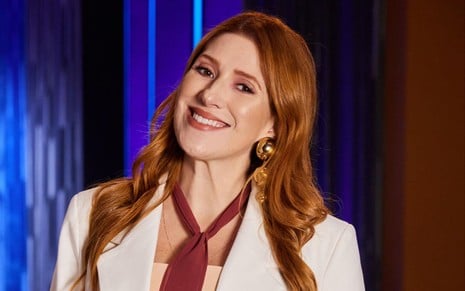 Mulher branca, de cabelos compridos e ruivos, usando blazer branco e gravata vermelha