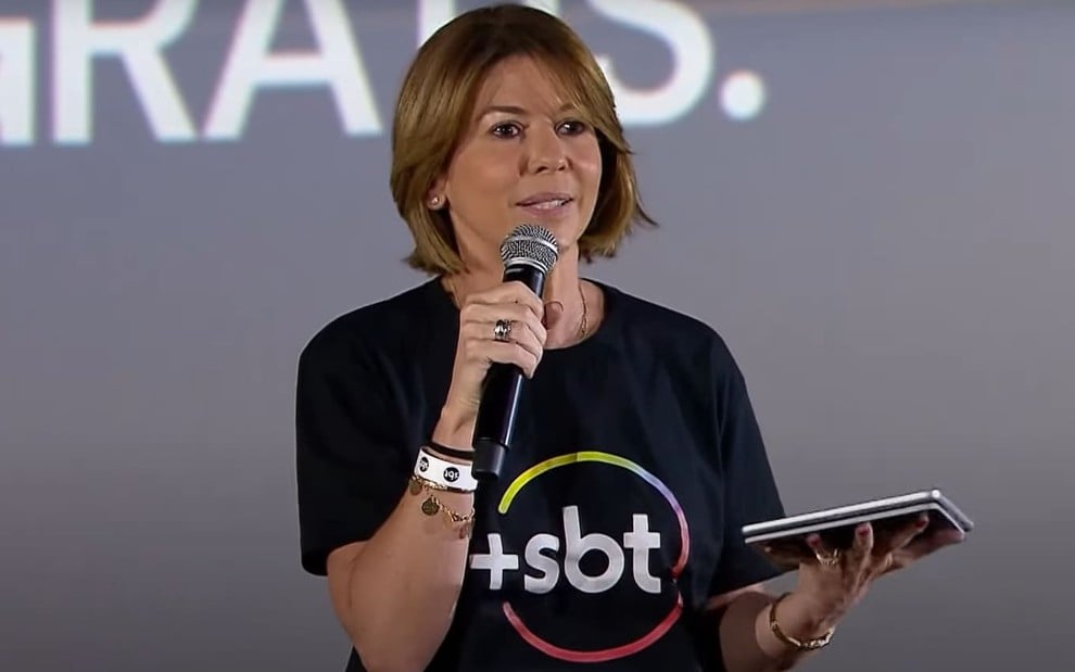 Daniela Beyruti está séria; ela segura um microfone e veste uma camiseta preta com o logo do +SBT