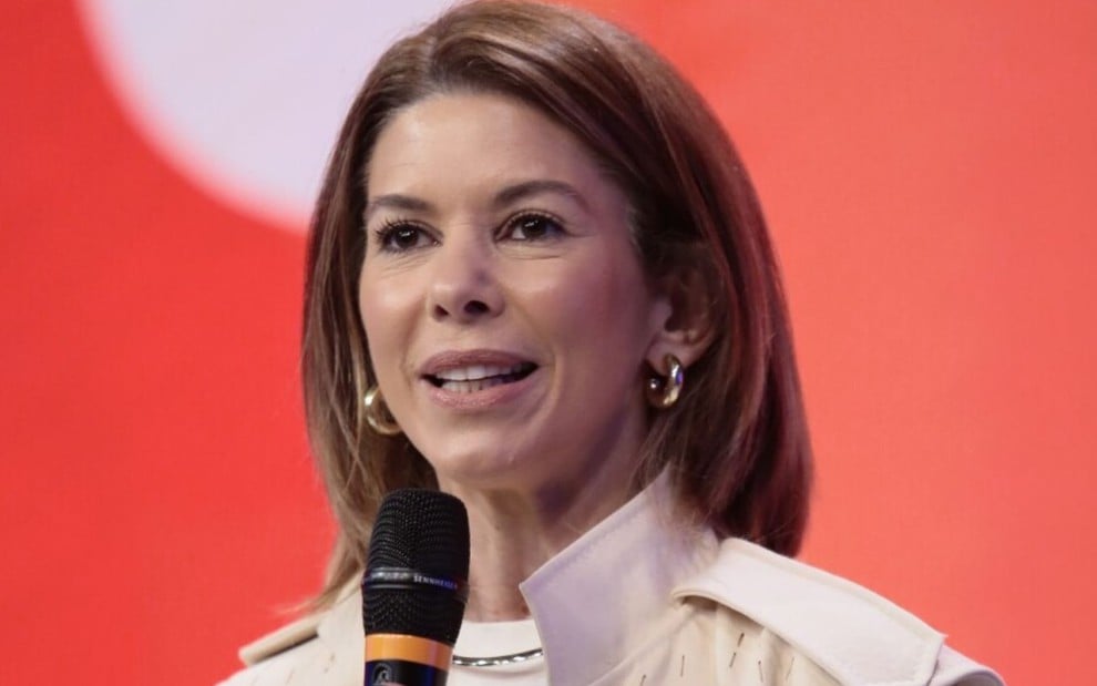 Daniela Beyruti fala em um microfone em frente a um fundo vermelho