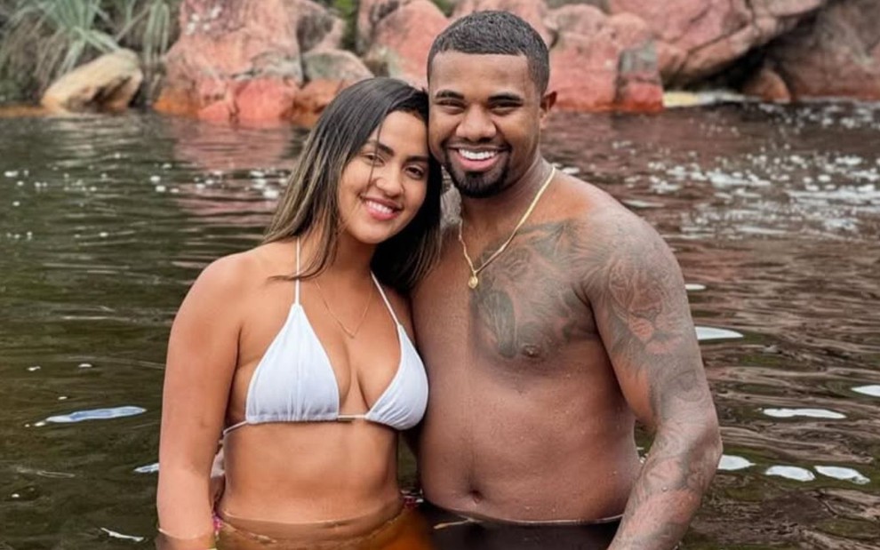 Adriana Paula e Davi Brito estão abraçados, com metade do corpo submerso em um rio e roupas de banho