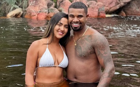 Adriana Paula e Davi Brito estão abraçados, com metade do corpo submerso em um rio e roupas de banho