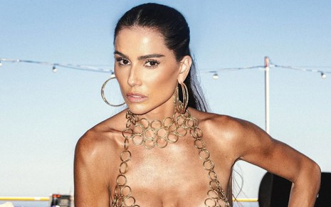 Deborah Secco posando com feição séria, usando um top de correntes, em frente a um céu azulado