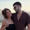Um casal posa sorridente diante de uma paisagem urbana com montanhas ao fundo;