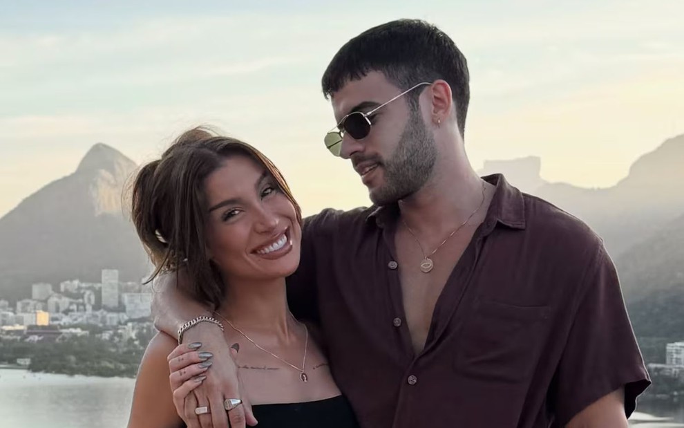 Um casal posa sorridente diante de uma paisagem urbana com montanhas ao fundo;