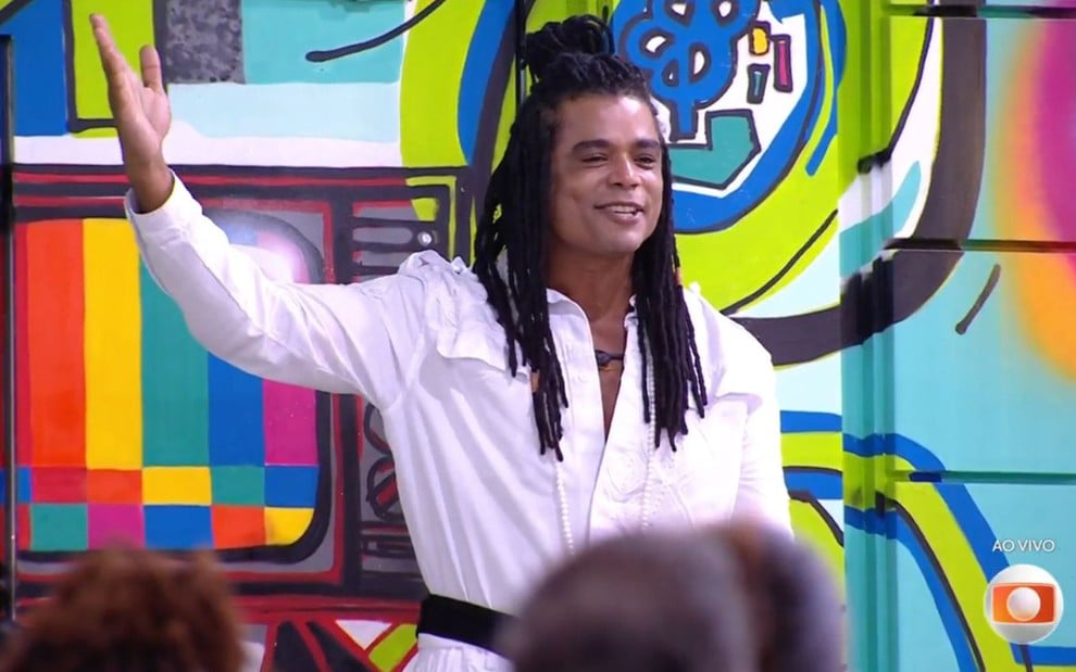 Diogo Almeida se despede de participantes no BBB 25; ele usa camisa branca, tem dreads no cabelo e está com o braço direito levantado