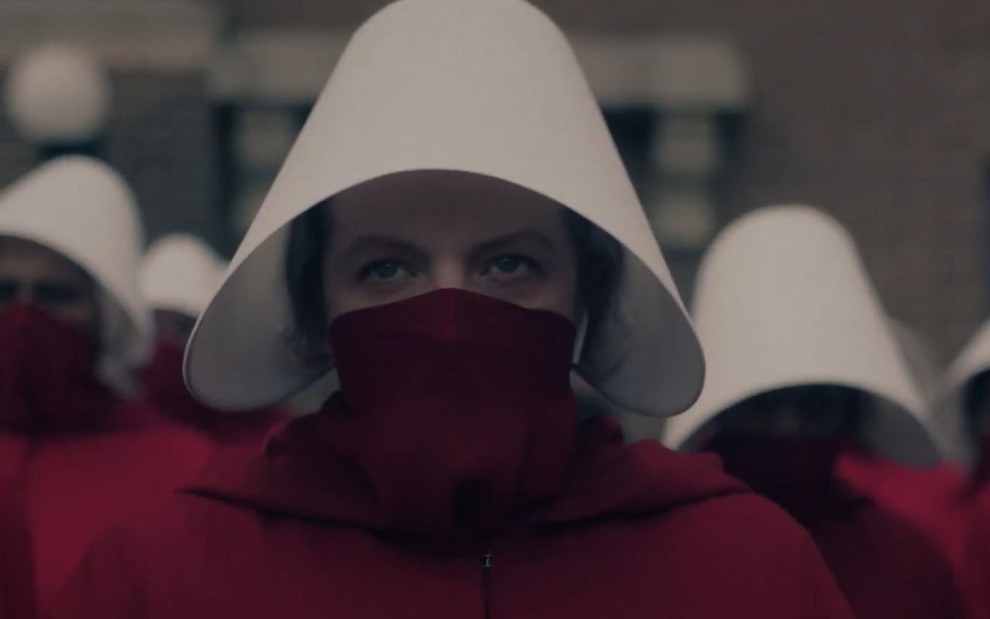 Elisabeth Moss usa uma roupa vermelha com chapéu branco; só é possível ver os olhos da atriz na veste típica das aias em The Handmaid's Tale