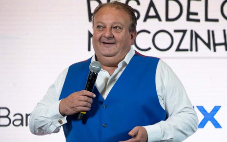 Erick Jacquin durante divulgação do reality Pesadelo na Cozinha, segurando microfone em frente a telão, sorrindo